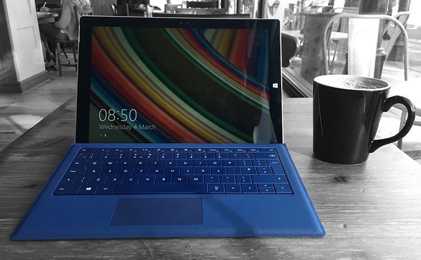 A Microsoft Surface Pro 3 with a blue keyboard. It is on a table in a coffee shop. There is a coffee mug next to it.